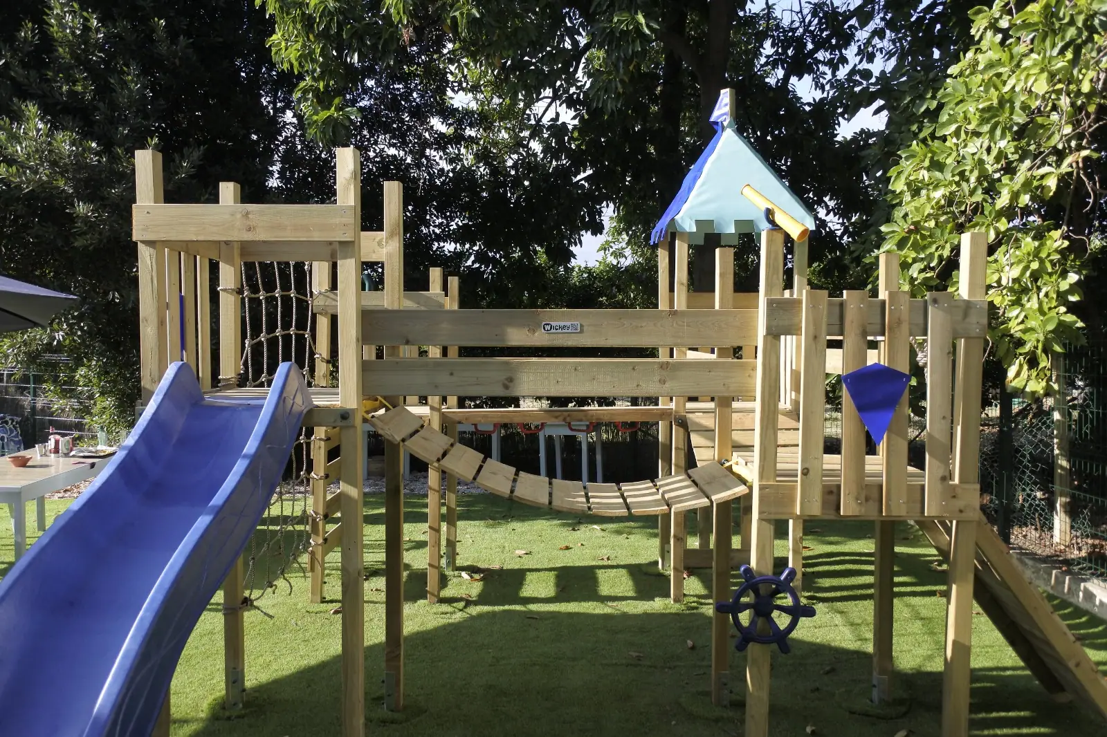 Zazoo's big wooden play castle with a blue slide, rope bridge and ship's wheel on astro turf