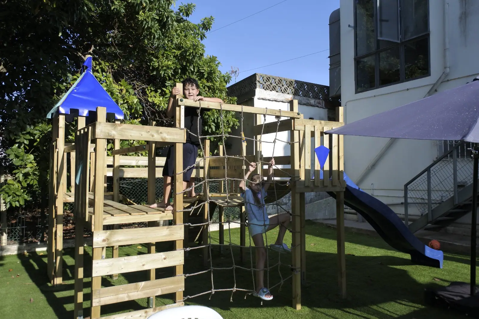 Kids climbing the wooden play frame beside the blue outdoor slide at Zazoo