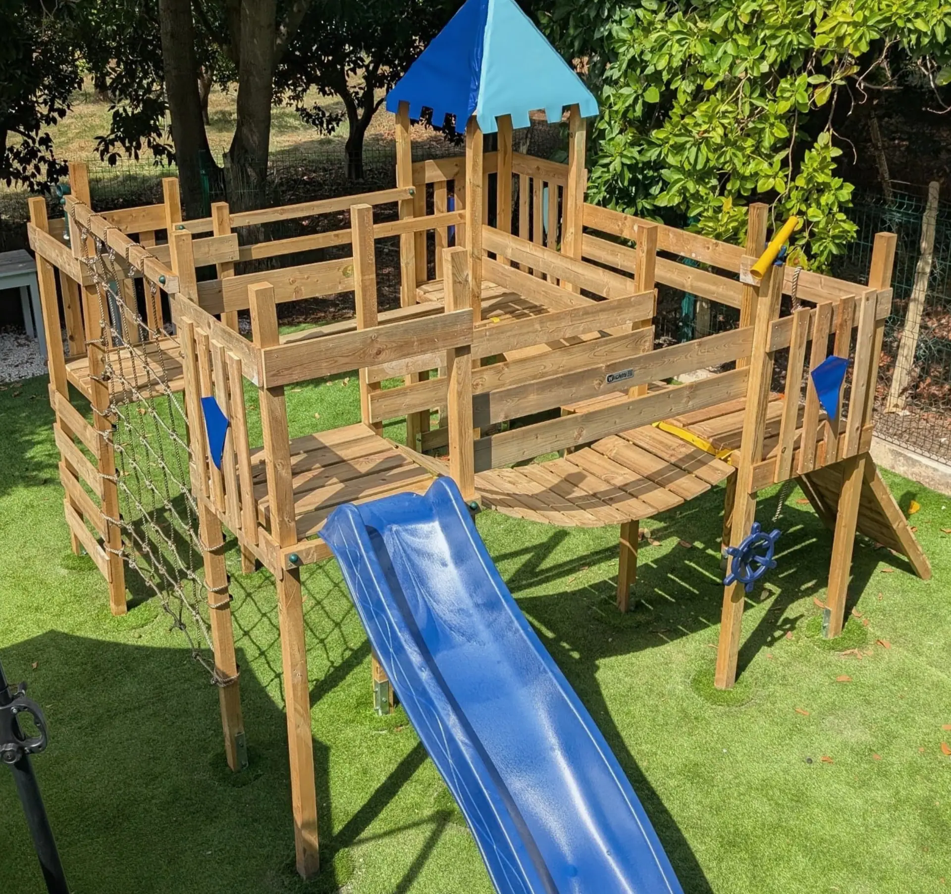 Zazoo's outdoor wooden play castle with a blue slide and climbing net on astro turf